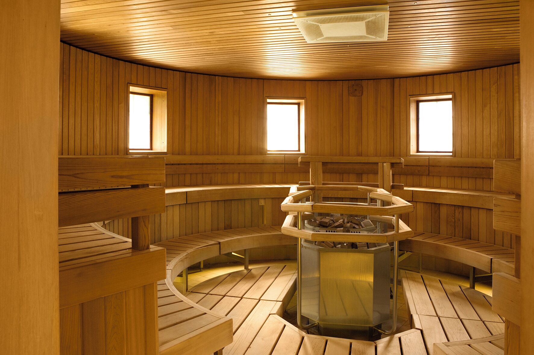 A sauna with wooden walls and benches, a central heating unit, and three windows.