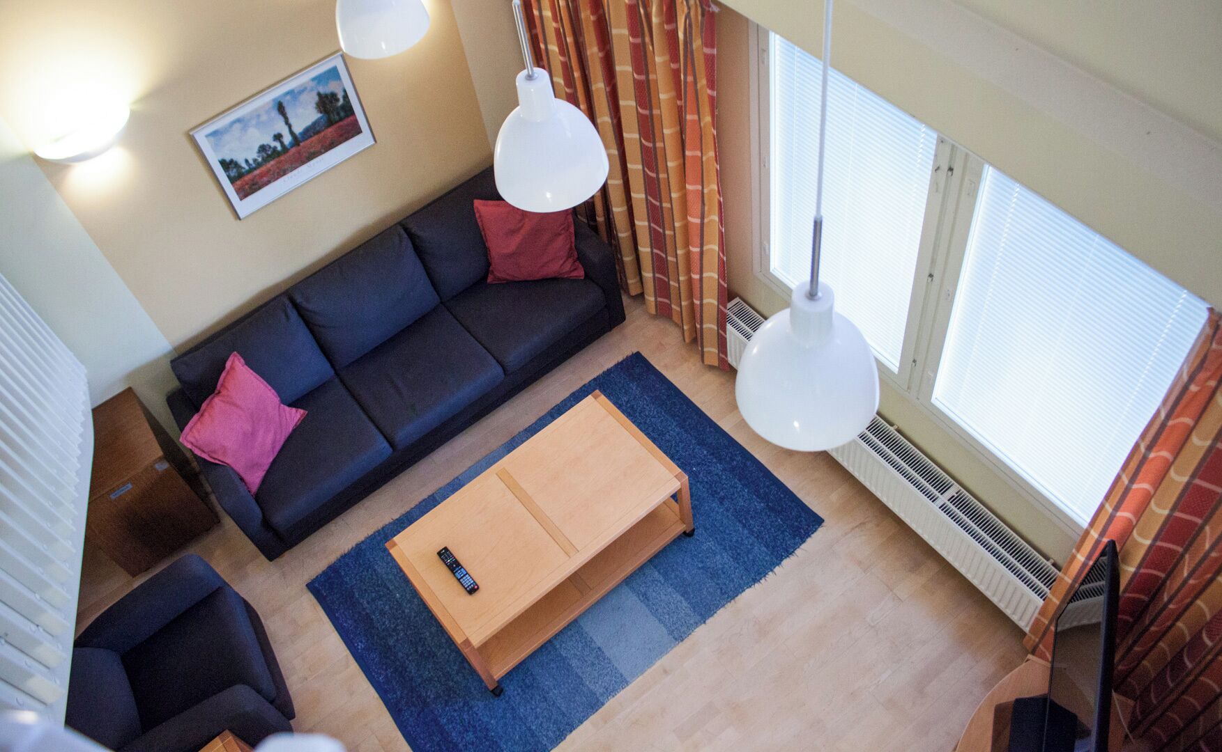 A modern living room with a dark sofa, a wooden coffee table, and a blue rug. There are two pendant lights, a framed picture on the wall, and a window with curtains.