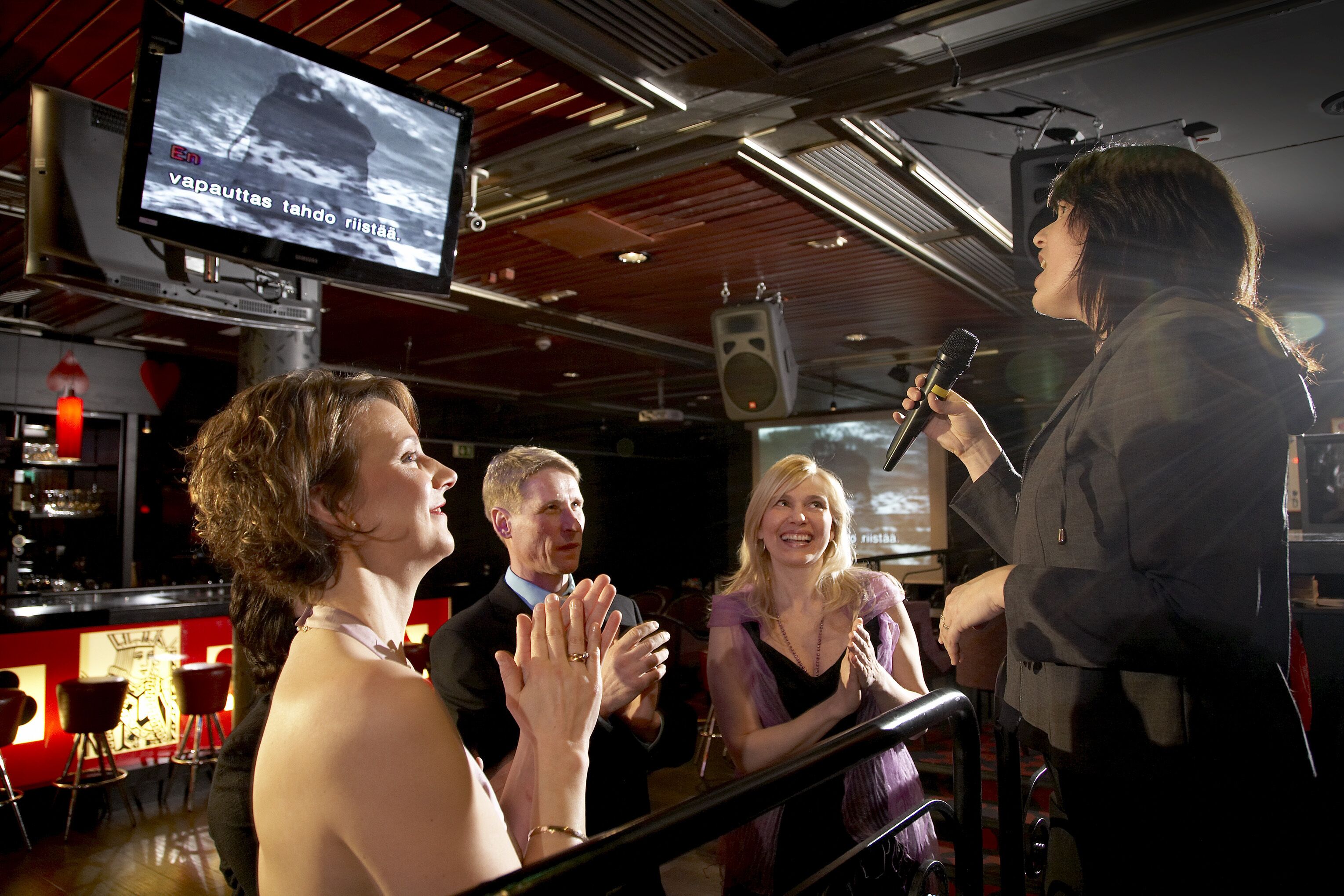 A bar with a television screen displaying a news channel, a bar counter, and a group of people standing and clapping.