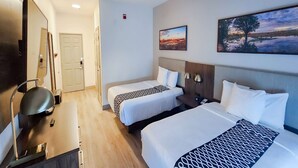 Una camera d'albergo con due letti, una scrivania, una sedia, una lampada e due quadri appesi al muro.