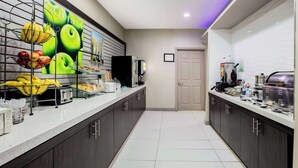 A hotel kitchen with a long counter, a fruit display, and a coffee station. - La Quinta Inn & Suites by Wyndham Dallas Mesquite (Mesquite)