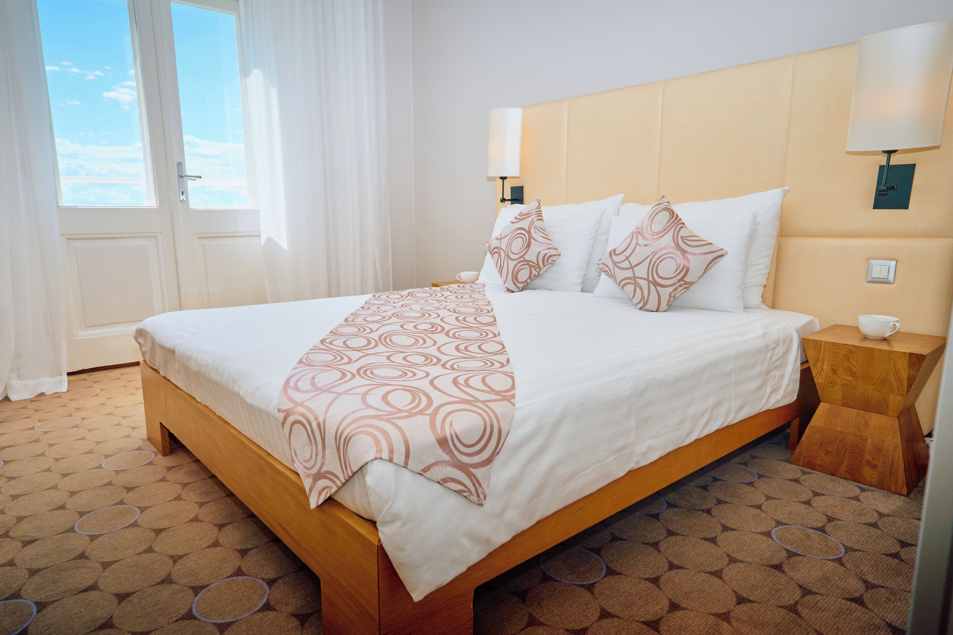 A hotel room with a large bed, a bedside table, a lamp, and a view of the sky through the window.
