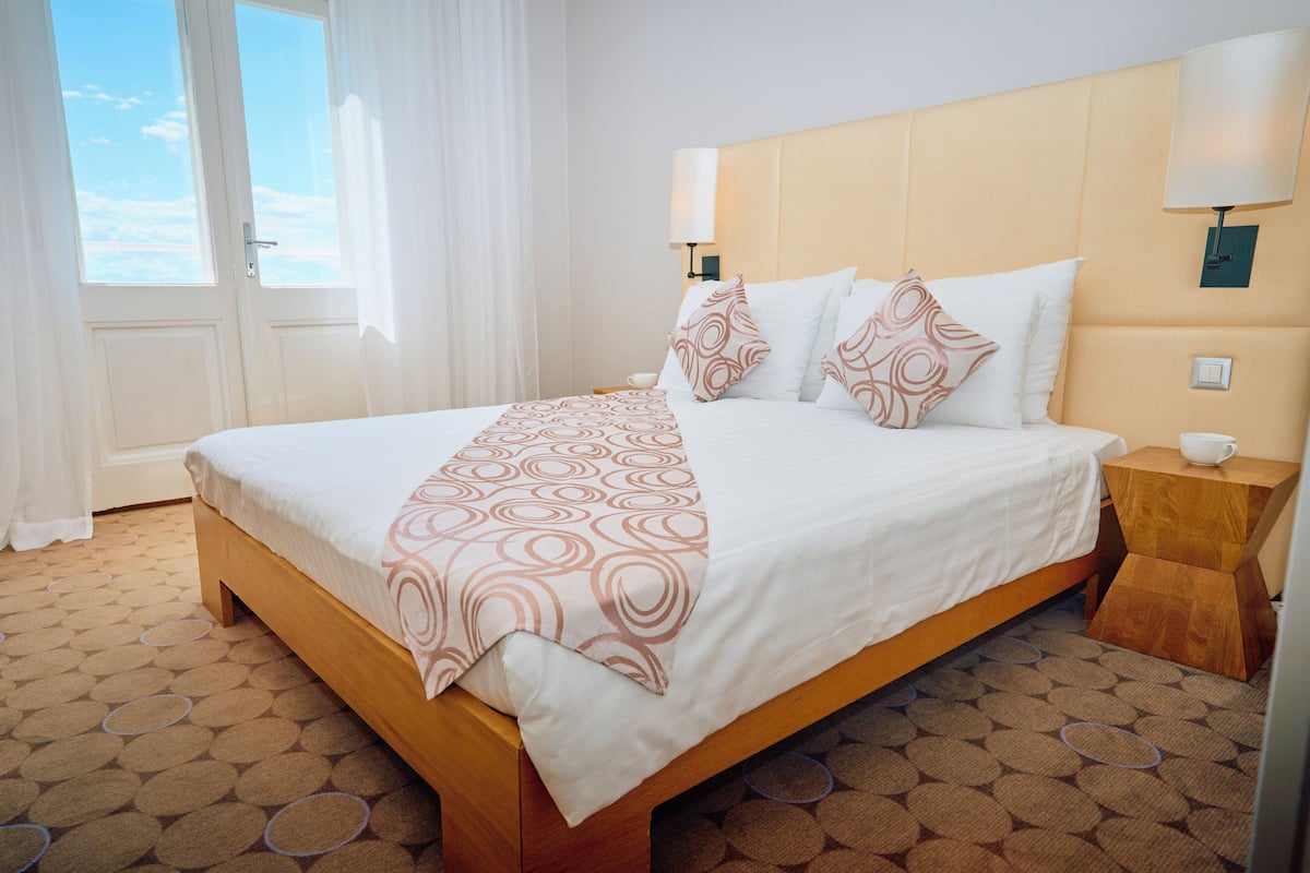 A hotel room with a large bed, a bedside table, a lamp, and a view of the sky through the window.