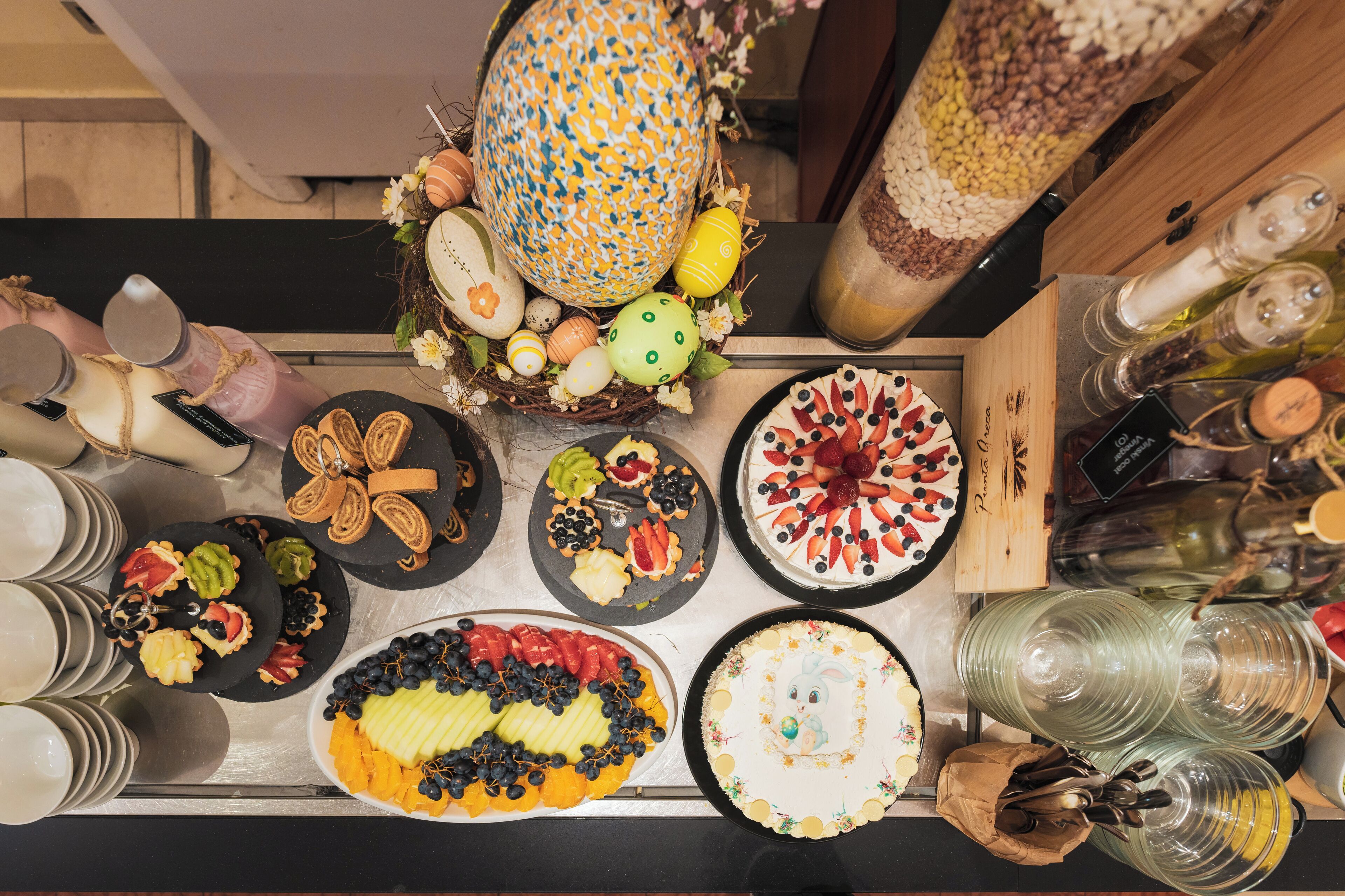 A table with various food items and decorations, including a large patterned vase, a basket of eggs, and several dishes with fruits and pastries.