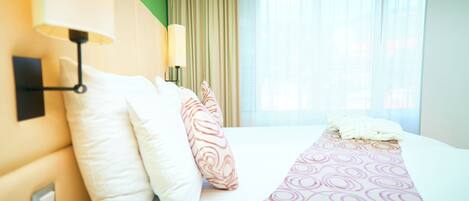 A hotel room with a bed, pillows, a lamp, and a window with curtains.