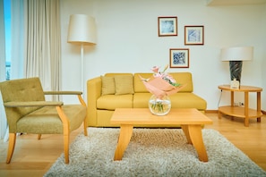 Sala de estar com sofá amarelo, mesa de centro de madeira, poltrona verde e tapete felpudo branco. Há duas luminárias, um quadro na parede e um vaso com flores sobre a mesa de centro.