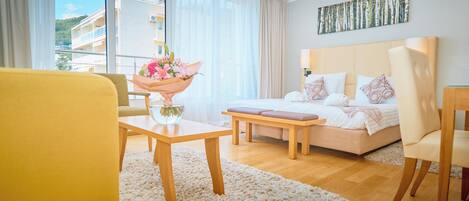 A modern hotel room with a bed, a sofa, a dining table, and a view of the outside.