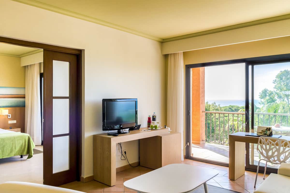 Habitación de hotel con cama, escritorio con televisor, balcón con vistas y zona de comedor.