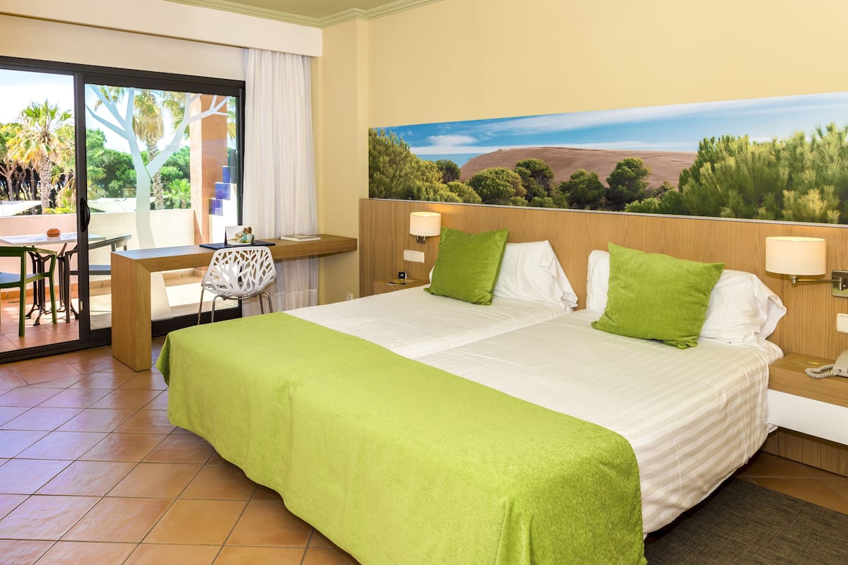 Habitación de hotel con cama, dos almohadas verdes, mesita de noche con lámpara, escritorio con silla y vista a árboles por la ventana.
