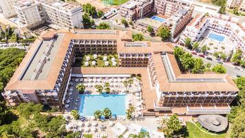 An aerial view of a hotel with a central swimming pool, multiple balconies, and surrounding greenery.
