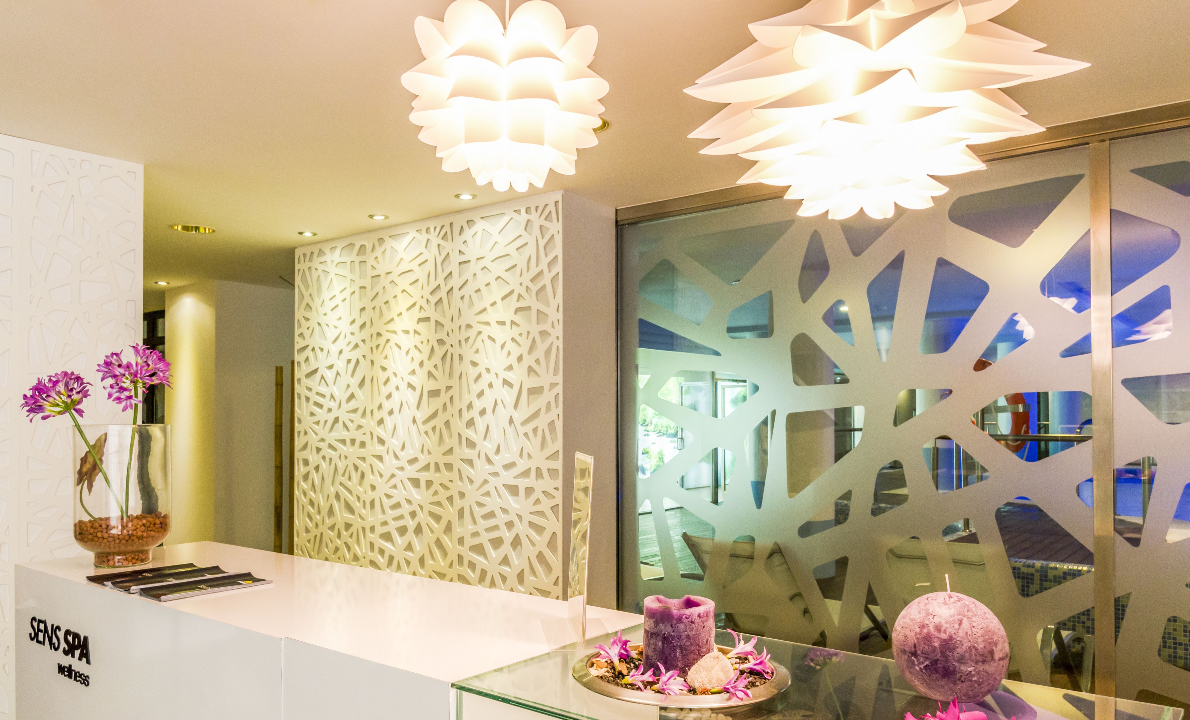 Un comptoir d’accueil avec un plan de travail blanc, un panneau décoratif orné de motifs géométriques découpés, et une vitrine en verre contenant une bougie violette et des fleurs.