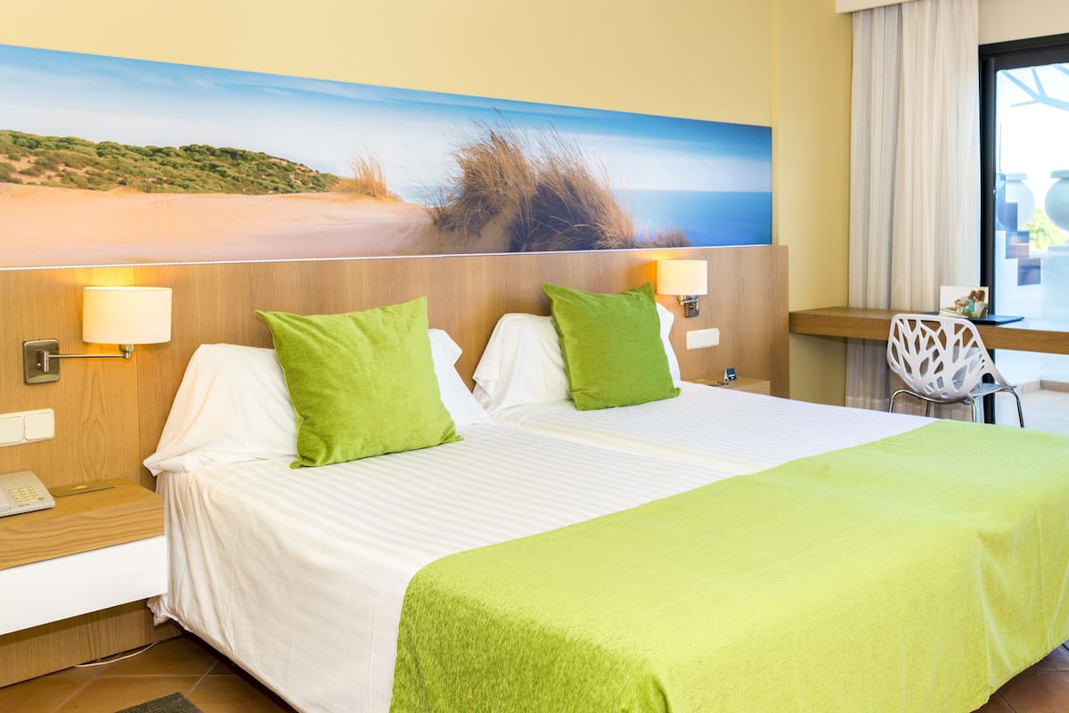 Habitación de hotel con una cama, almohadas verdes, una mesita de noche, una lámpara y un cuadro con una escena de playa.