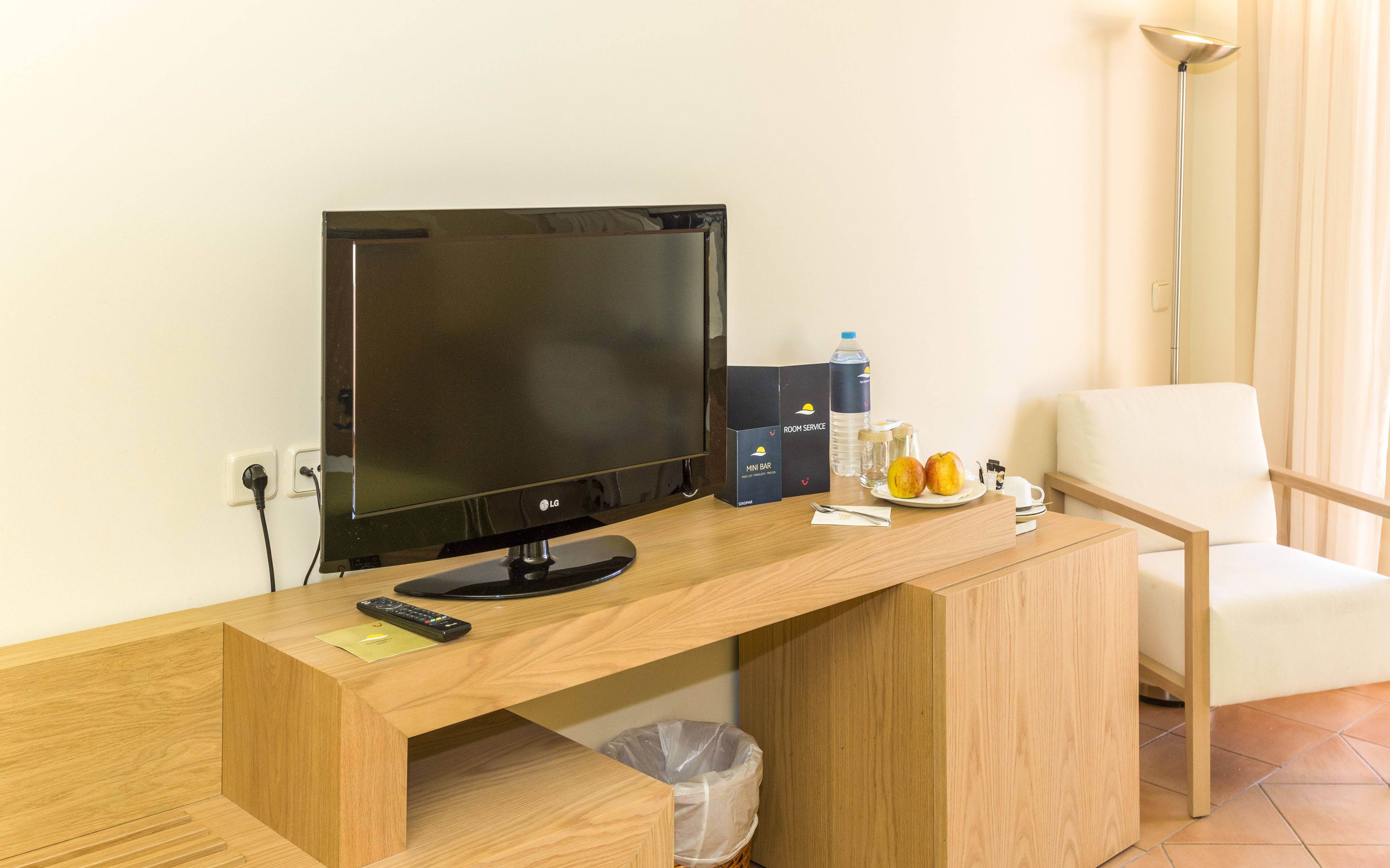 A hotel room with a wooden desk, a flat-screen TV, a mini bar, a chair, and a trash bin.