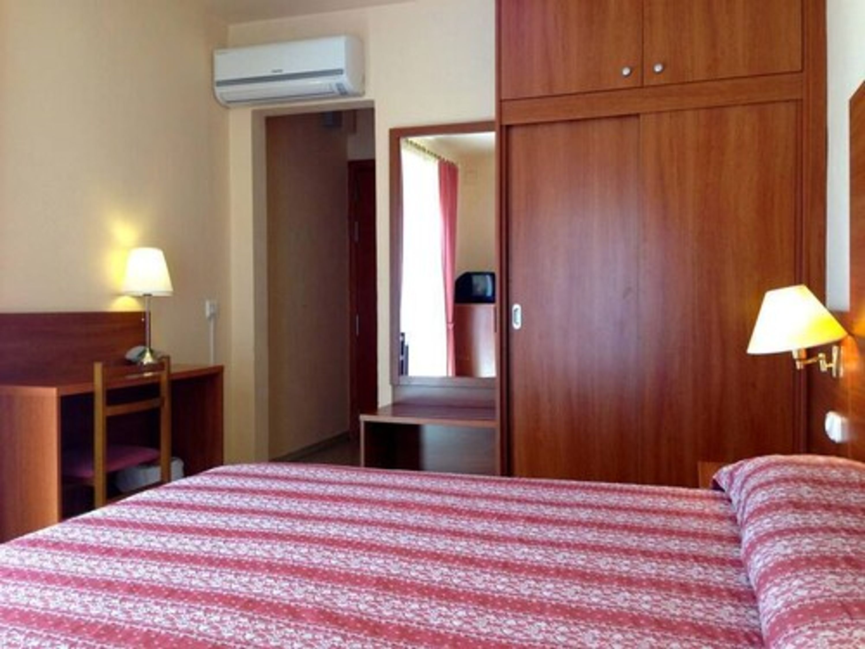 A hotel room with a bed, two bedside lamps, a wooden wardrobe, and an air conditioning unit.