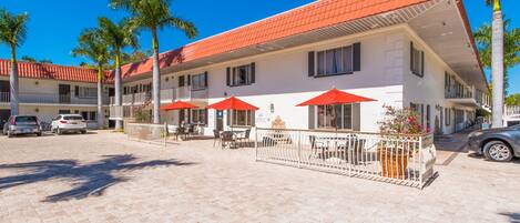 A hotel with a red-tiled roof, outdoor seating with red umbrellas, and palm trees.