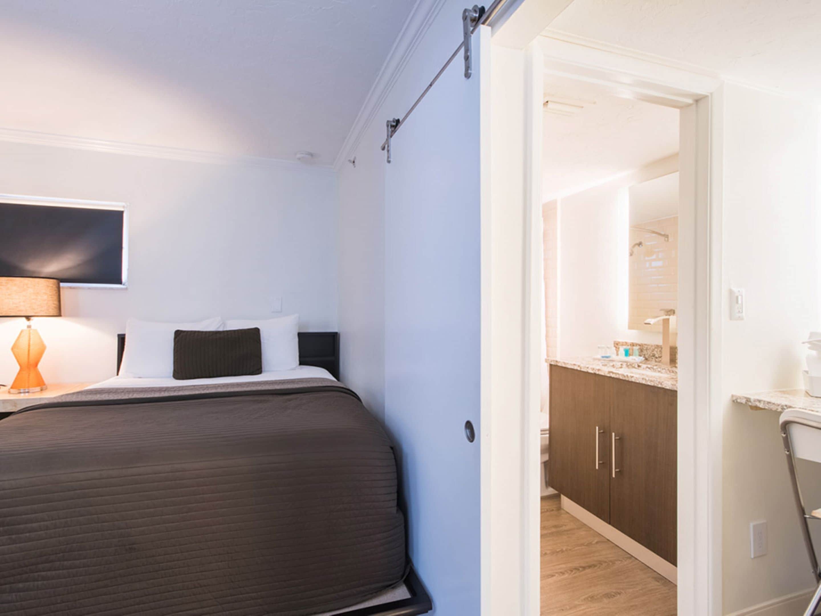 A hotel room with a bed, a nightstand with a lamp, and a view into the bathroom.