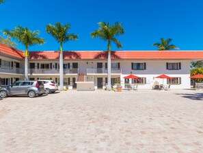 A hotel with a red-tiled roof, multiple balconies, and palm trees. There are cars parked in front and outdoor seating with red umbrellas. - Hibiscus Suites - Gateway to Siesta Key (Sarasota)