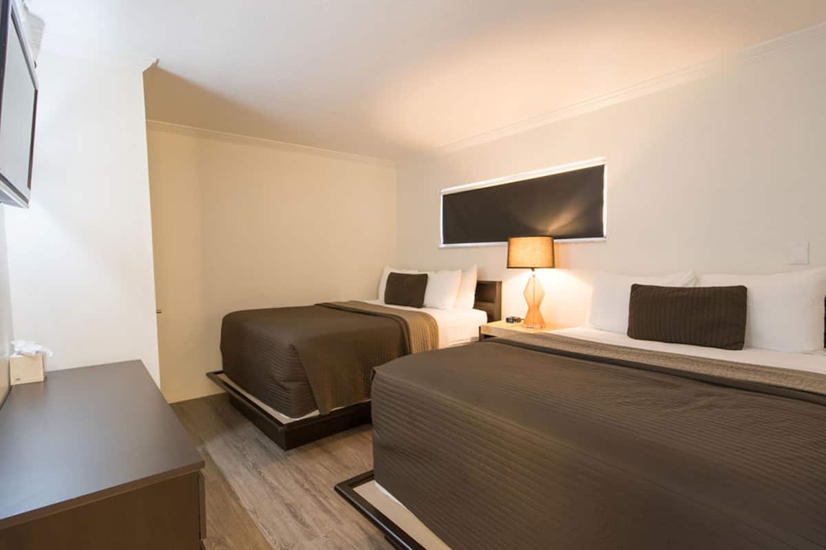 A hotel room with two beds, a desk, a flat-screen TV mounted on the wall, and a bedside table with a lamp.