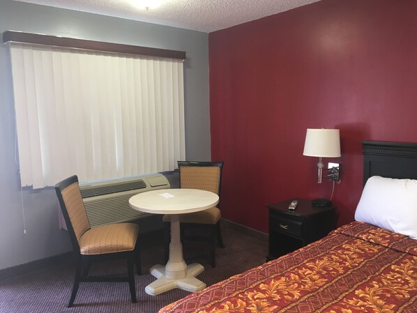 A hotel room with a bed, a small table with two chairs, a nightstand with a lamp, and a window with blinds.