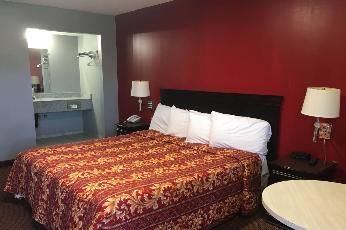 A hotel room with a large bed, a round table, a nightstand with a lamp, and a mirror.