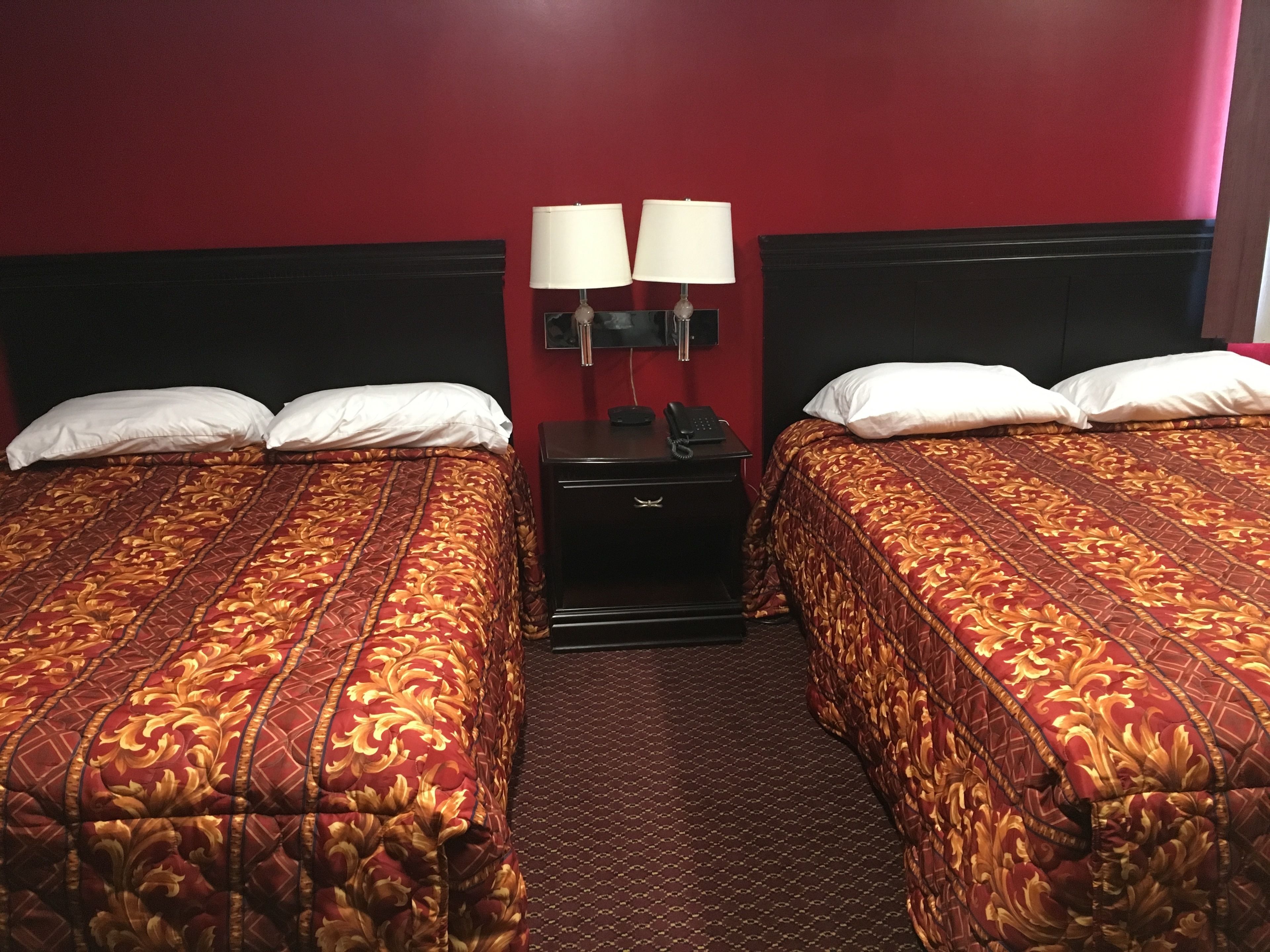 A hotel room with two beds, each with a red and gold patterned bedspread, a black nightstand with a lamp, and a red wall.
