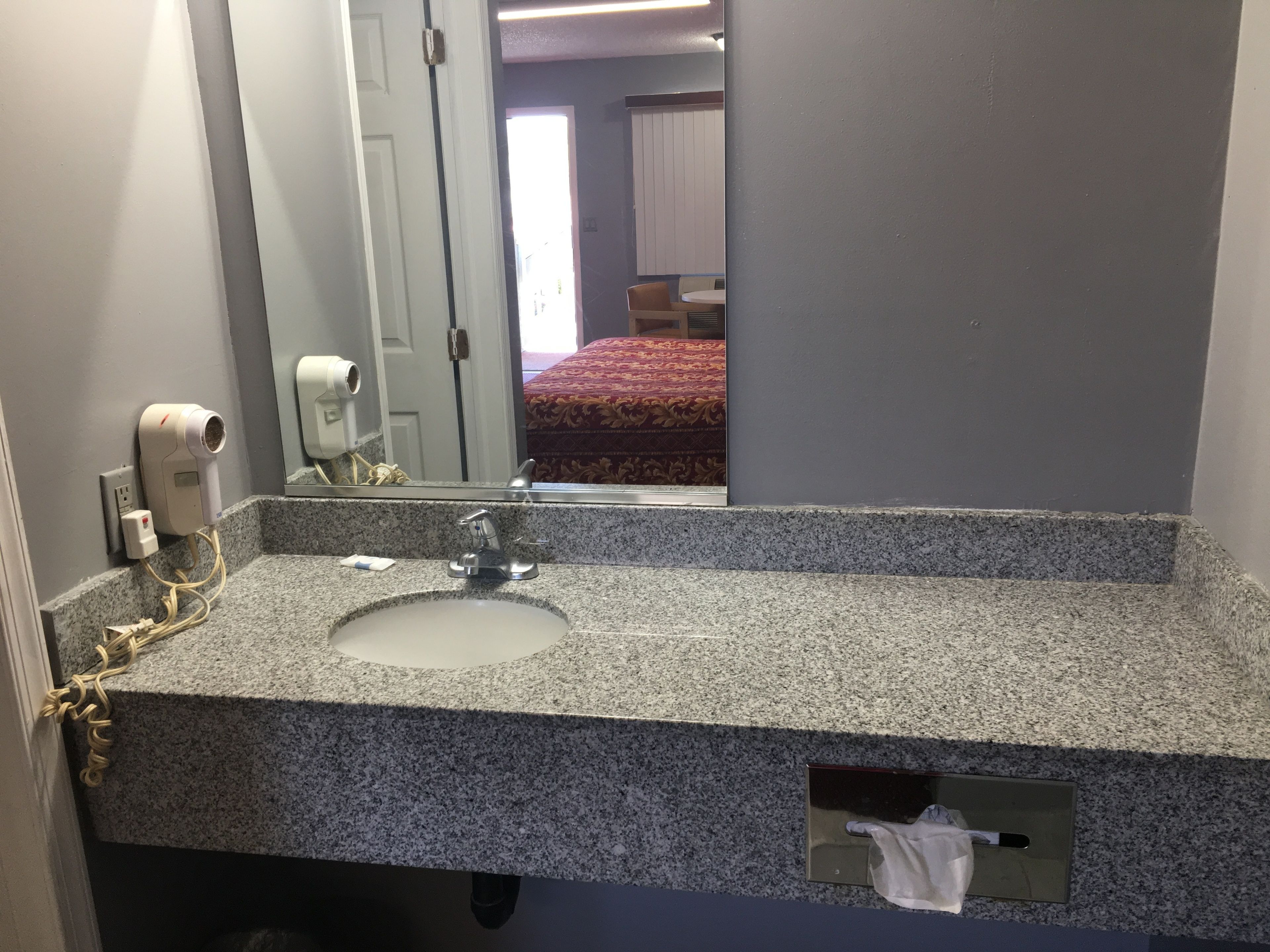Een badkamer met een granieten aanrecht, een wastafel, een spiegel, een haardroger en een zeepdispenser.