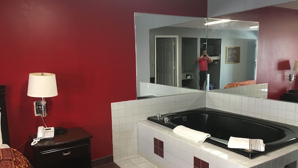 Habitación de hotel con un espejo grande, una mesita de noche negra con lámpara, alfombra roja, bañera blanca con azulejos y pared roja.
