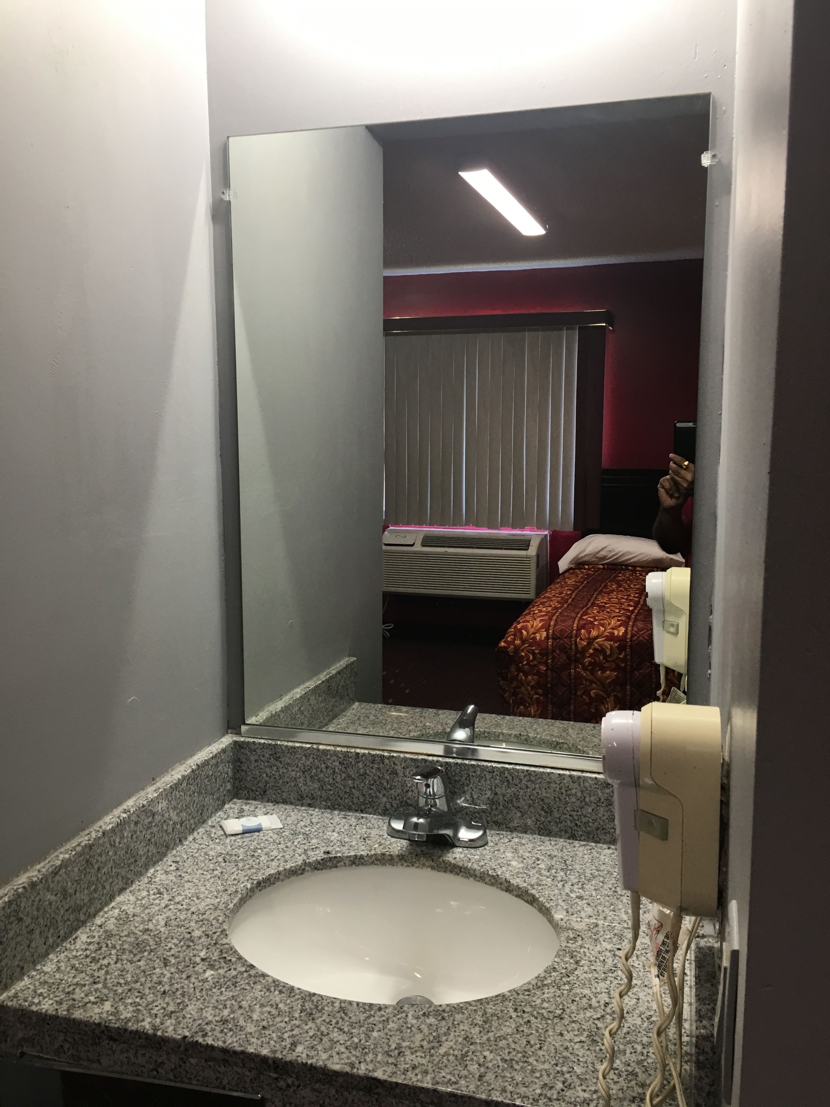 Un baño con lavamanos de granito, un espejo, un secador de manos y una cama con una colcha de cuadros rojos y blancos.