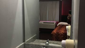 A bathroom with a granite sink, a mirror, a hand dryer, and a bed with a red and white patterned bedspread.
