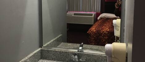 Un baño con lavamanos de granito, un espejo, un secador de manos y una cama con una colcha de cuadros rojos y blancos.