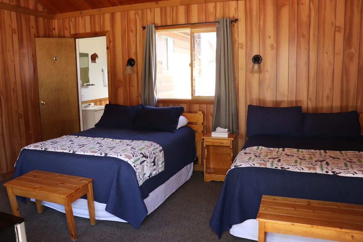 A wooden cabin room with two beds, a small table, a wooden bench, a mirror, and a window with curtains.