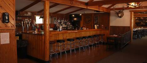 A bar area with wooden beams, a bar counter, stools, and a mounted deer head.