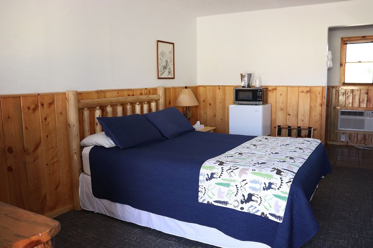 A bedroom with a wooden headboard, a bed with a blue bedspread, a bedside lamp, a small white refrigerator, and a microwave on a wooden shelf.
