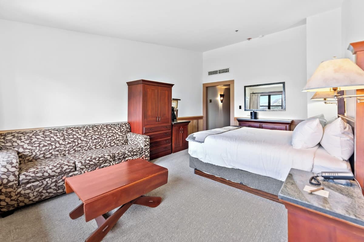 A room with a patterned sofa, a wooden table, a bed with white bedding, a wooden cabinet, and a mirror on the wall.