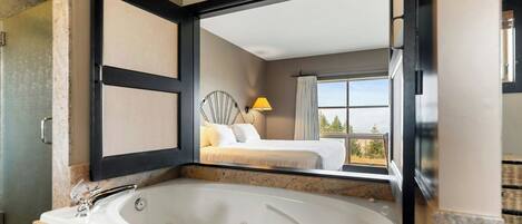 A modern bathroom with a large bathtub, a mirror reflecting a bedroom with a bed and a window, and a view of trees.
