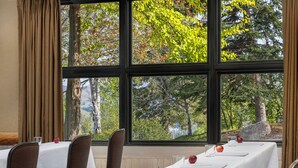 A dining area with a long table set for a meal, large windows with a view of trees and hills, and brown upholstered chairs. - Sun Mountain Lodge (Winthrop)