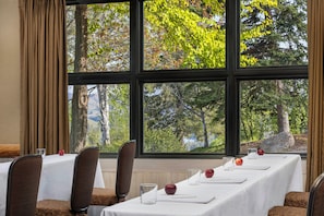 Ein Essbereich mit einem langen Tisch, der für eine Mahlzeit gedeckt ist, großen Fenstern mit Blick auf Bäume und Hügel sowie braunen, gepolsterten Stühlen.