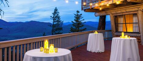 A deck with round tables, string lights, and candles, overlooking a mountainous landscape.
