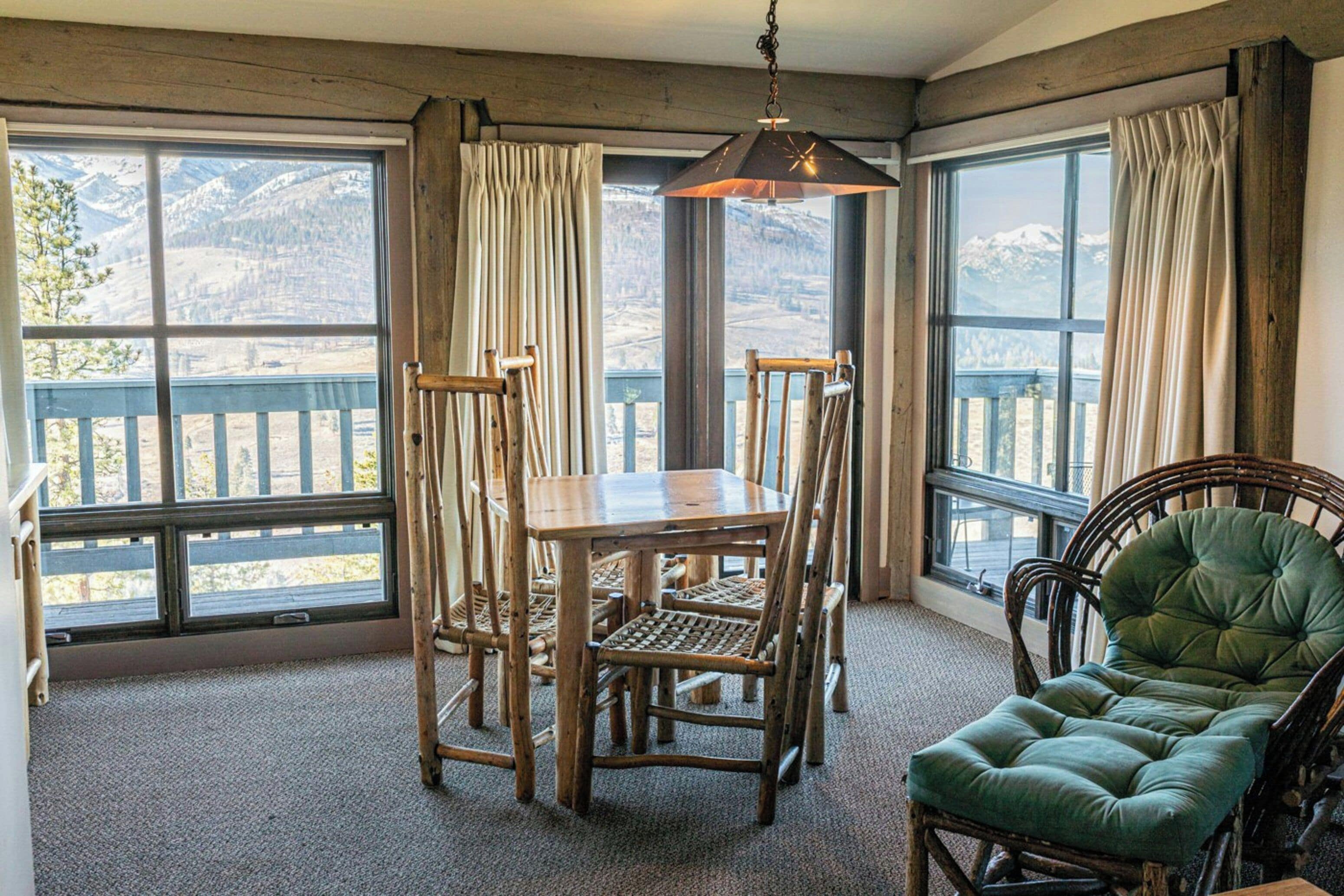 A room with a wooden dining table and chairs, a green upholstered chair, and large windows with curtains.