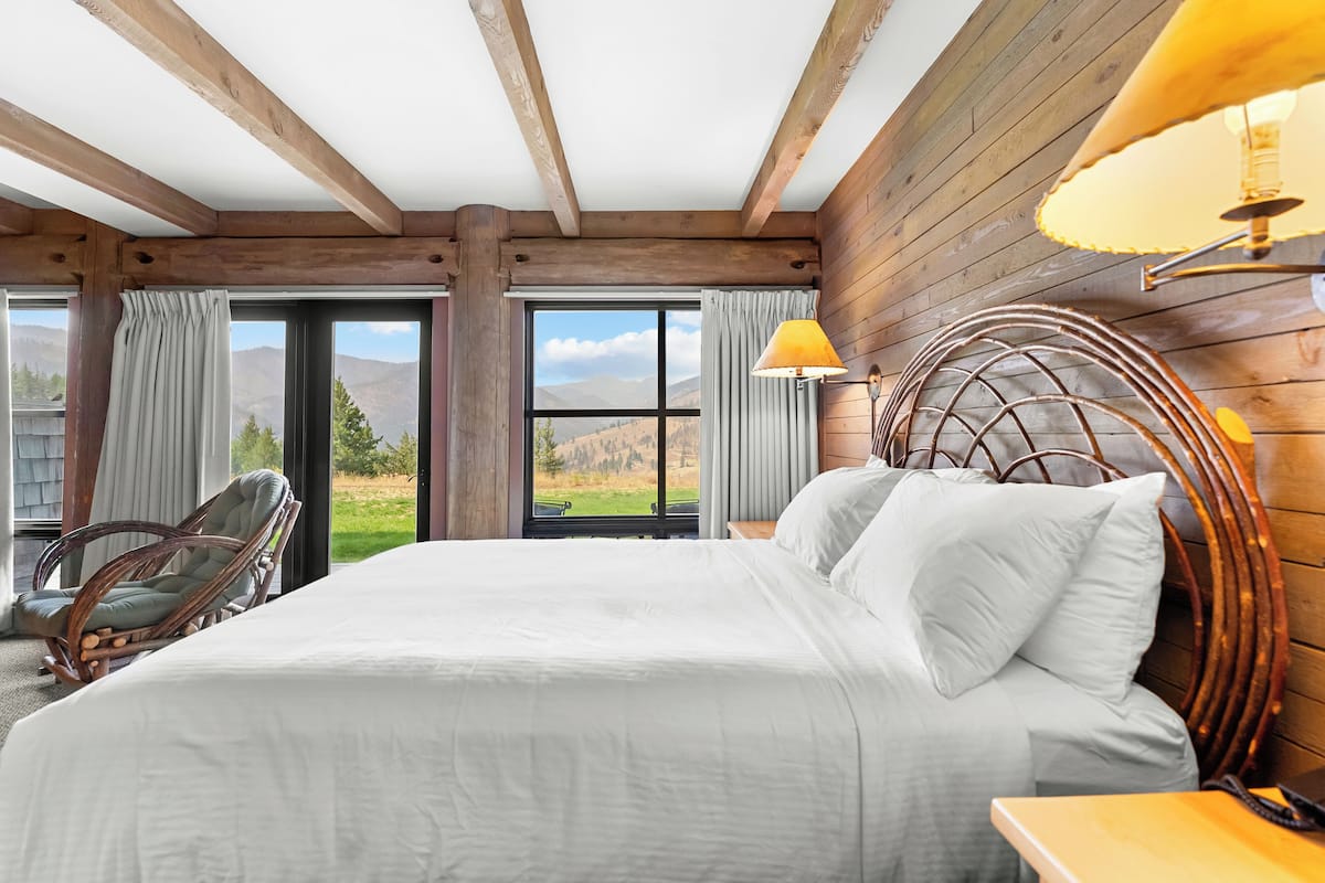 A bedroom with a large bed, wooden ceiling beams, a view of the outdoors, a wicker chair, and a lamp.