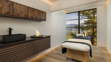 Quarto moderno com cama de madeira, mesa de cabeceira com velas e vista para uma paisagem montanhosa através de amplas janelas.