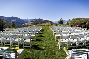Rows of white chairs with floral arrangements on a grassy aisle, set against a mountainous backdrop.