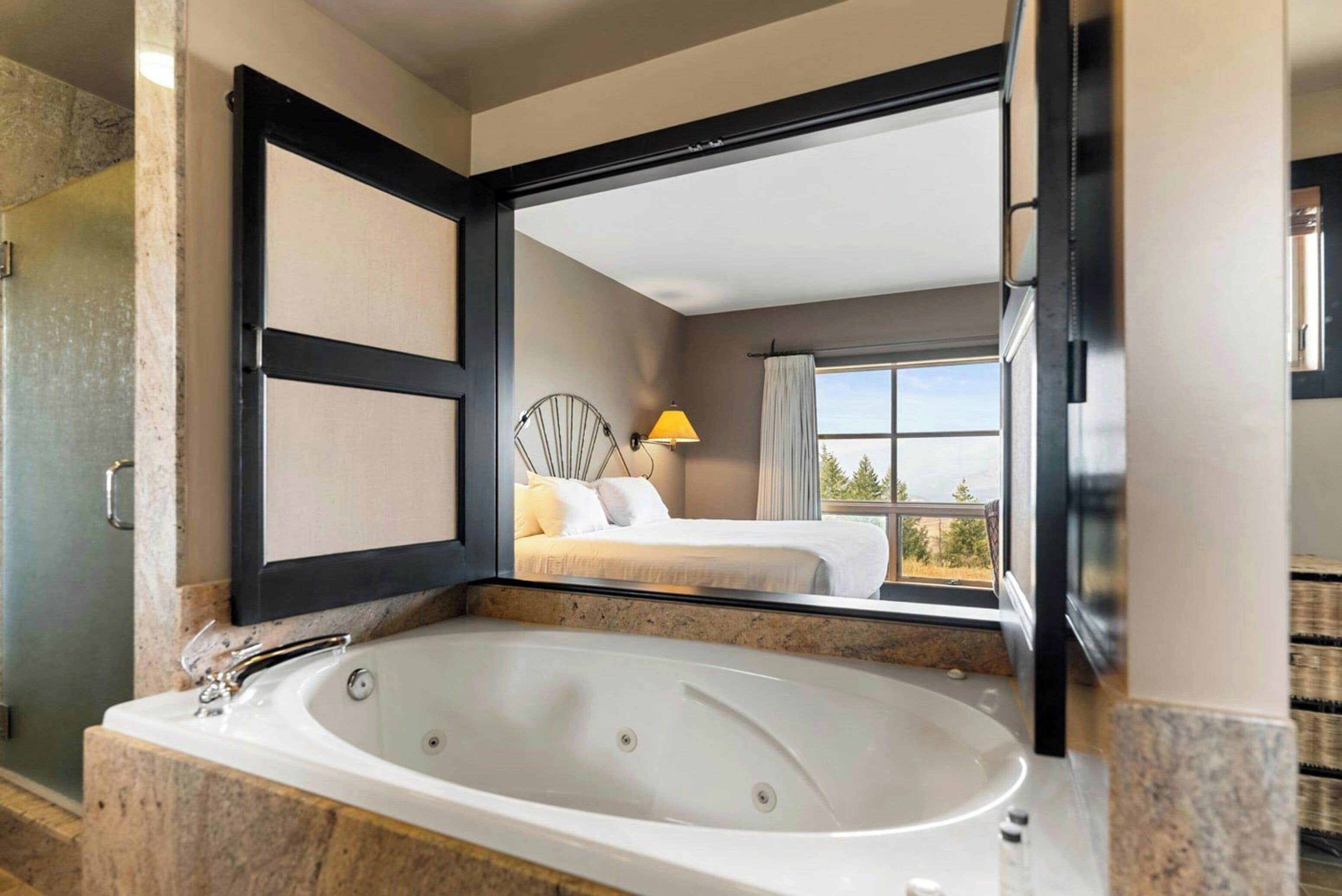 A modern bathroom with a large bathtub, a mirror reflecting a bedroom with a bed and a window, and a view of trees.