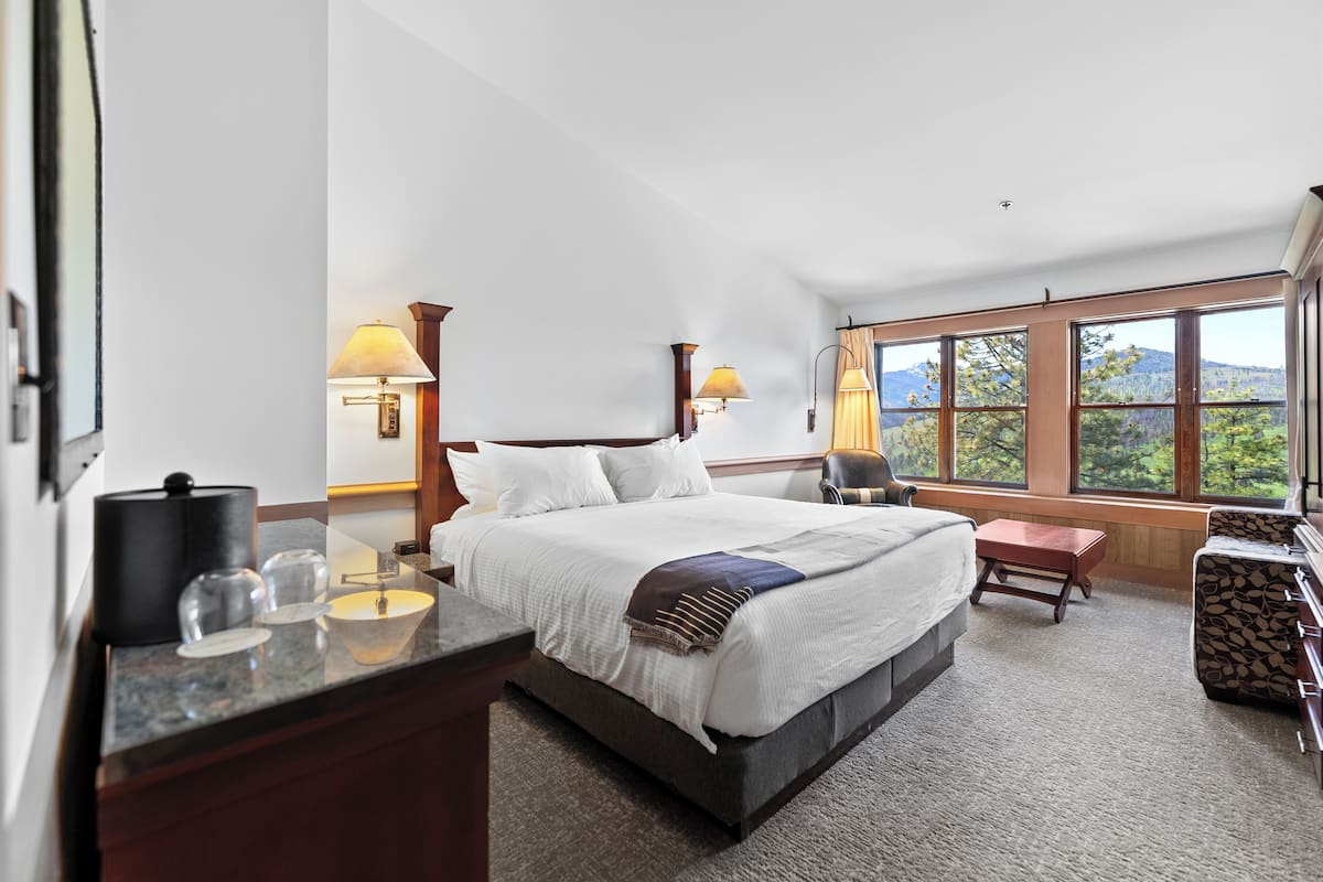 A spacious bedroom with a large bed, bedside lamps, a desk with a lamp, a chair, and a view of the outdoors through large windows.