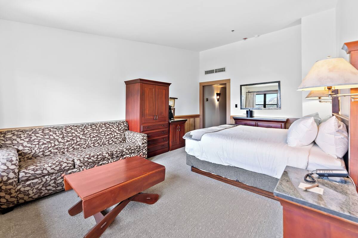 A room with a patterned sofa, a bed with white bedding, a wooden cabinet, and a desk with a lamp.