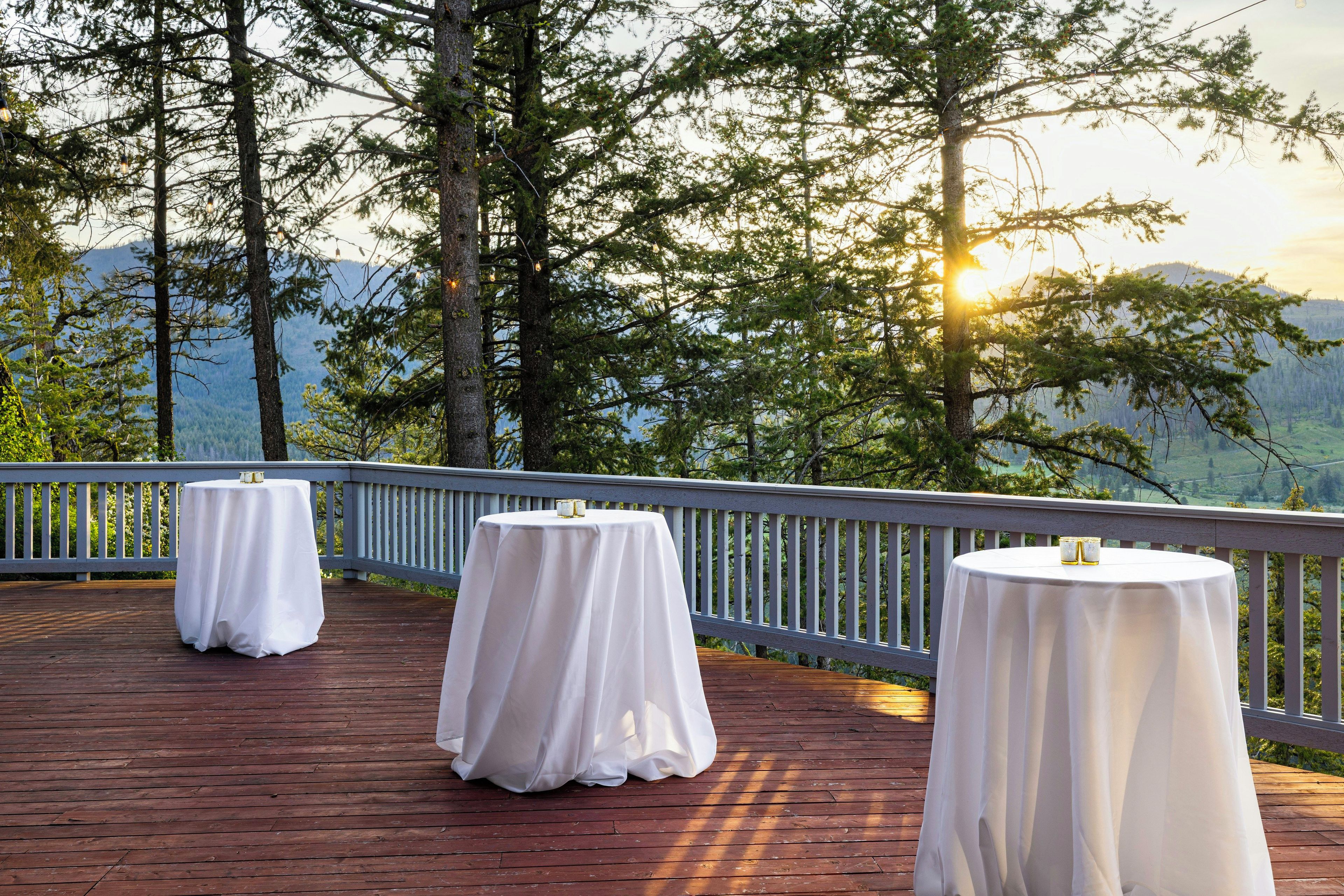 Une terrasse agrémentée de trois tables rondes recouvertes de nappes blanches, le tout mis en valeur par un décor de grands arbres et d’un paysage montagneux.