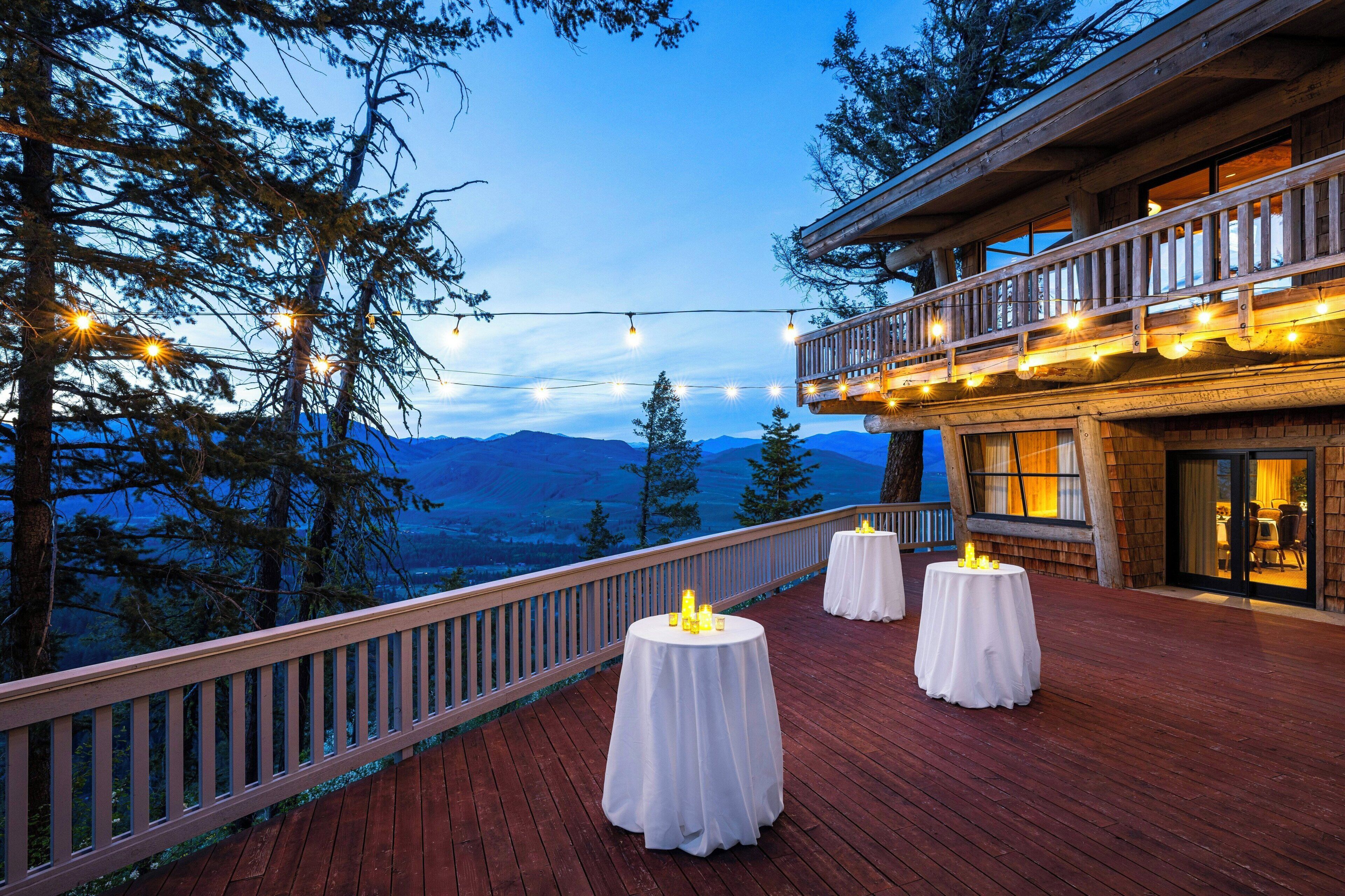 Une terrasse en bois, dotée de tables rondes et illuminée de guirlandes lumineuses, qui offre une vue sur un paysage montagneux.