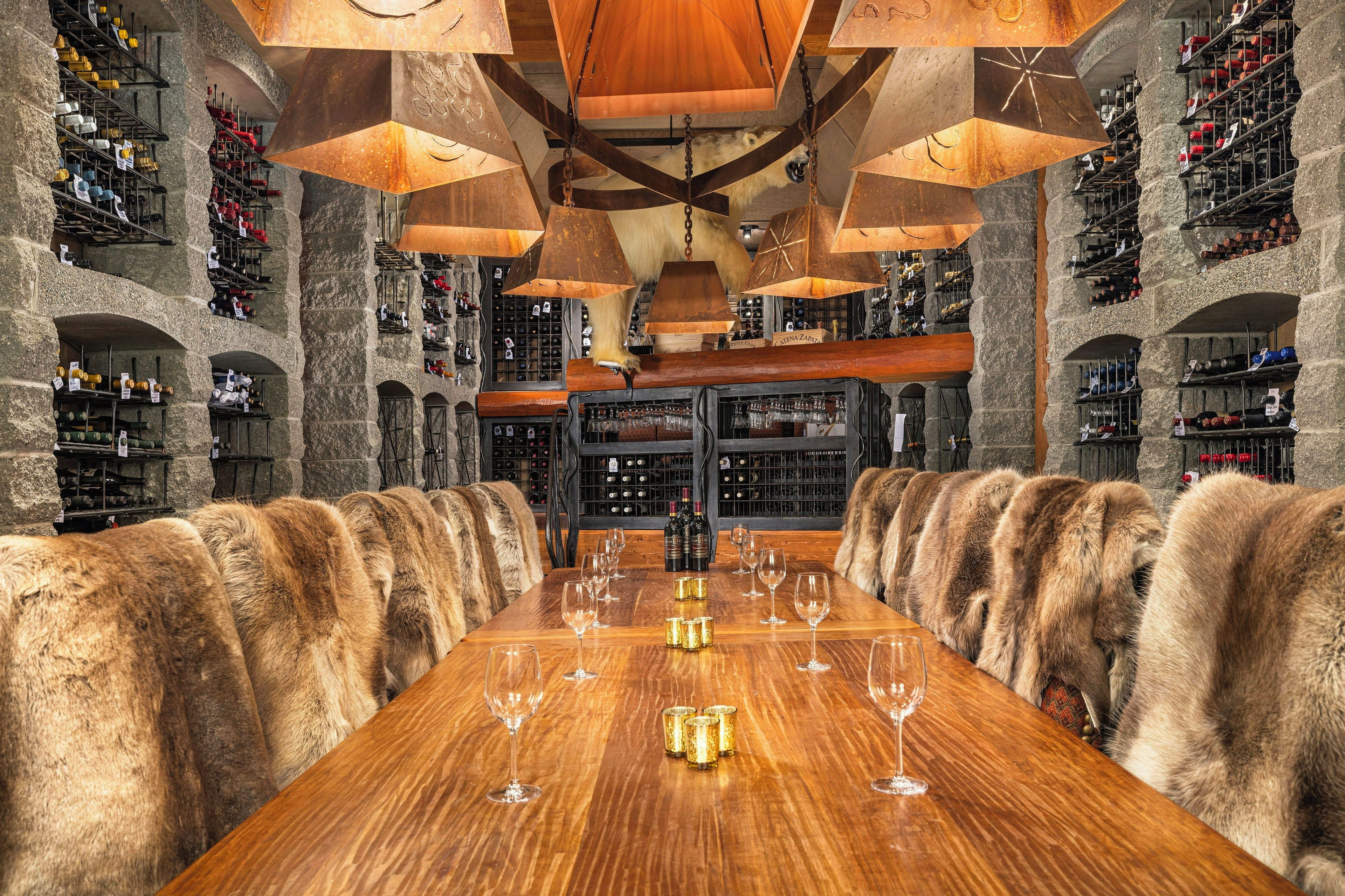 A spacious wine cellar with a long wooden table set for dining, surrounded by stone walls and wine racks.