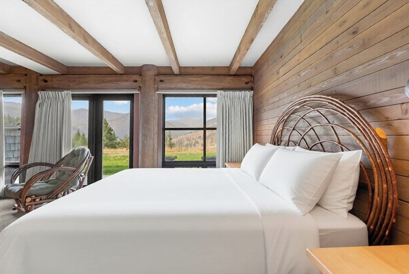 Une chambre avec un grand lit, des poutres en bois, une tête de lit en osier et une vue sur l’extérieur.