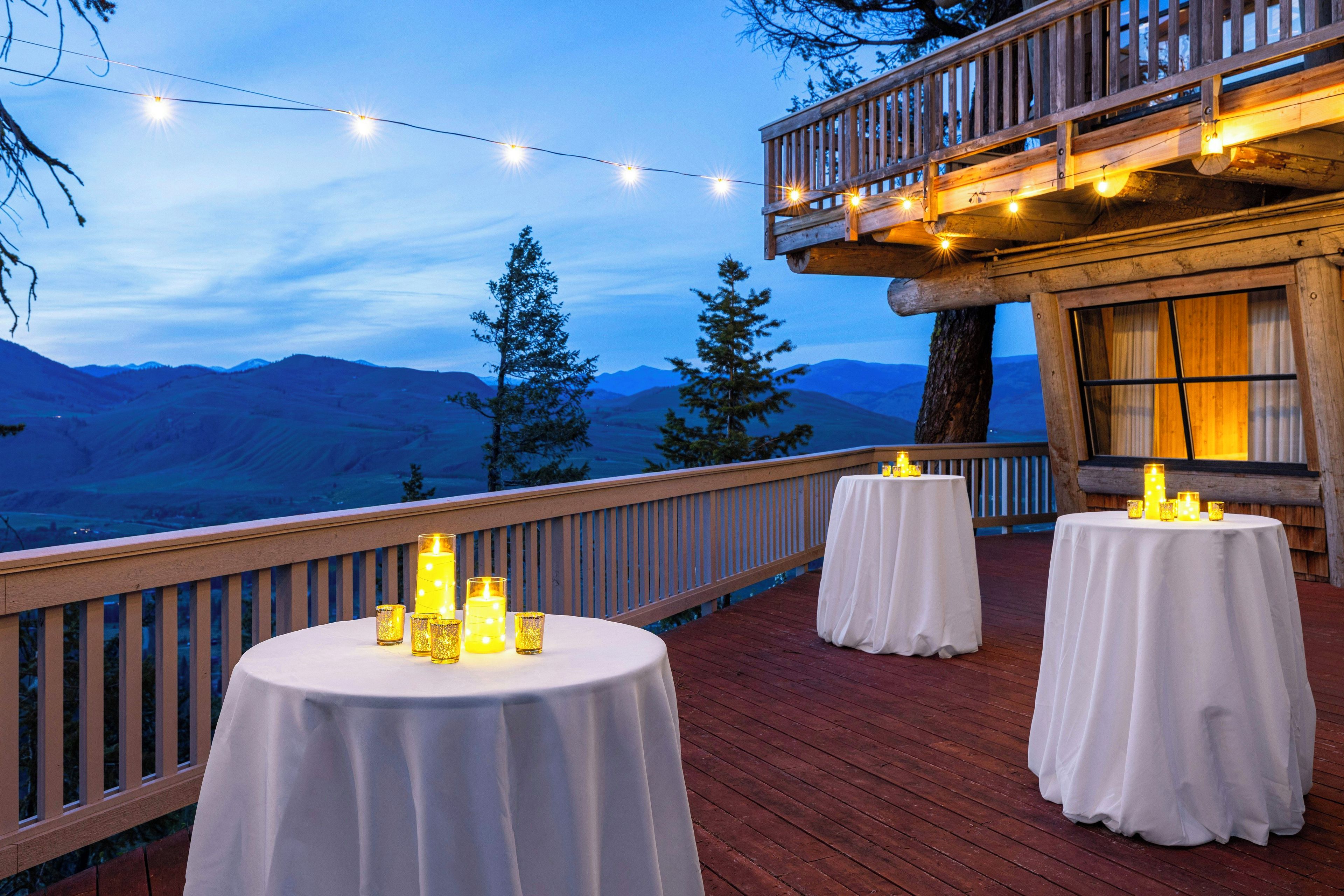 A deck with round tables, string lights, and candles, overlooking a mountainous landscape.
